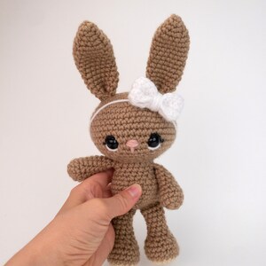 May include: A brown crocheted bunny with a white bow headband and black eyes. The bunny is standing on a white surface.