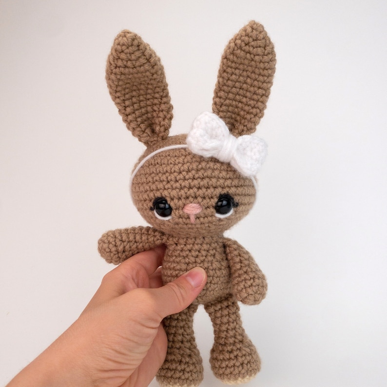 May include: A brown crocheted bunny with a white bow headband and black eyes. The bunny is standing on a white surface.