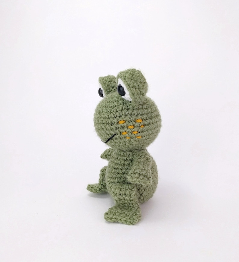 May include: A green crocheted frog toy with yellow spots on its face. The frog has black eyes and a white outline around the eyes.