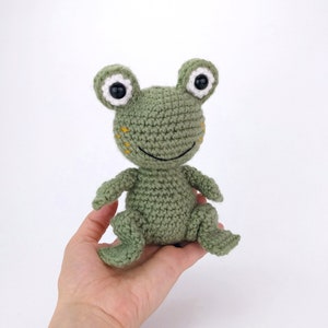 May include: A green crocheted frog toy with white eyes and a yellow smile. The frog is sitting on a hand.