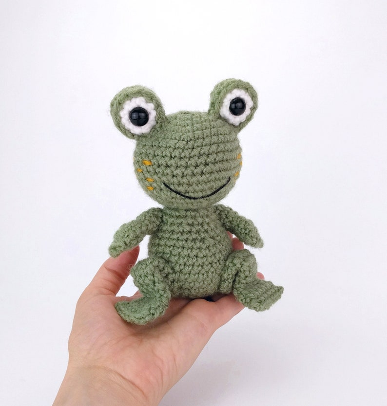 May include: A green crocheted frog toy with white eyes and a yellow smile. The frog is sitting on a hand.