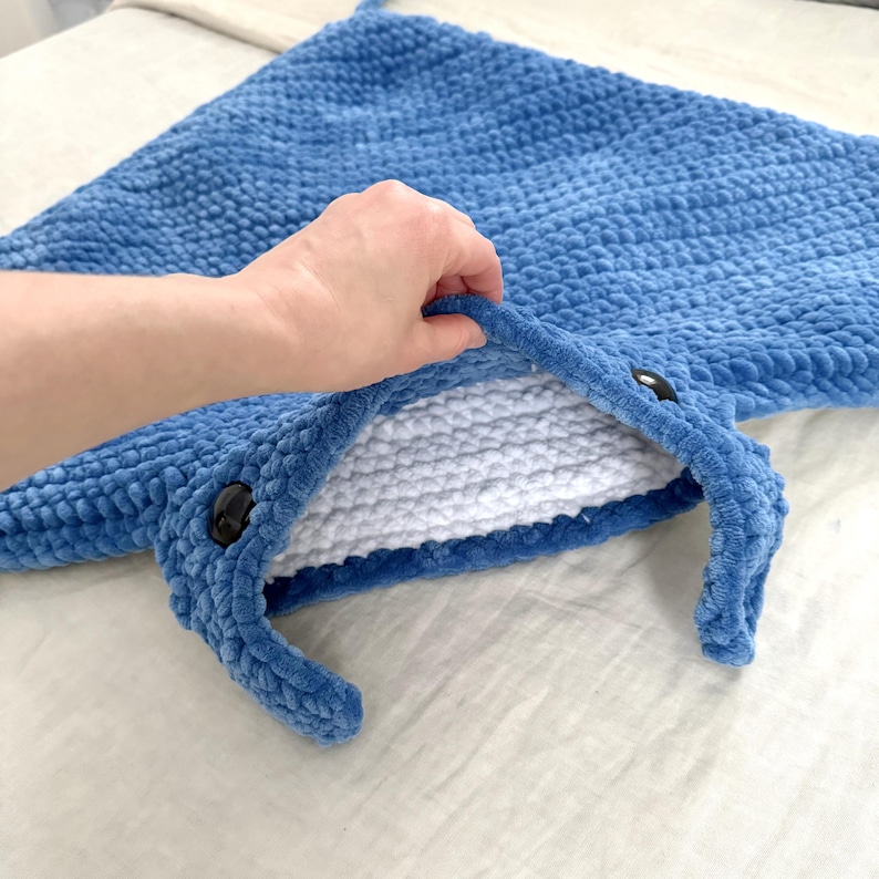May include: A blue crocheted manta ray blanket with white lining and black button eyes. The blanket is being held open to show the lining.