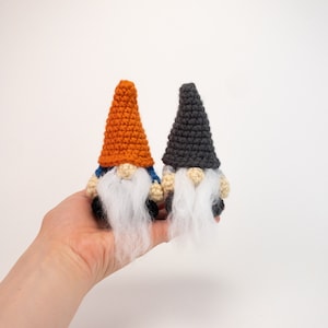 May include: Two crocheted gnomes, one with an orange hat and the other with a gray hat. Both gnomes have white beards and blue clothing.