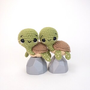 May include: Two crocheted green and brown turtle plushies with black eyes and smiling faces. The turtles are sitting on gray, geometric bases.