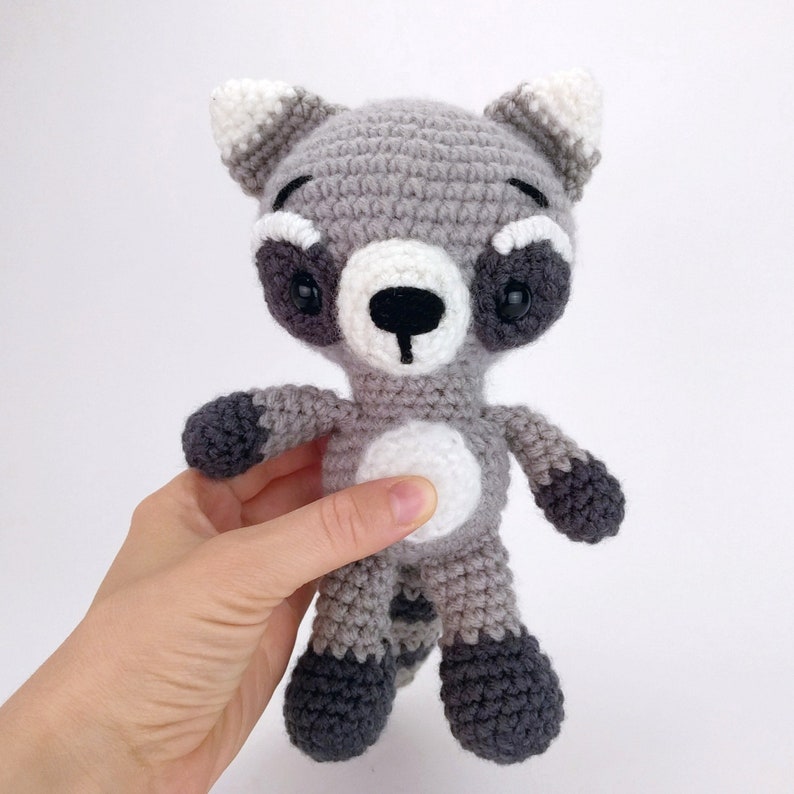 Puede incluir: Un juguete de peluche de mapache tejido a crochet con pelaje gris y blanco, ojos negros y nariz negra. El juguete est&aacute; de pie sobre una superficie blanca.