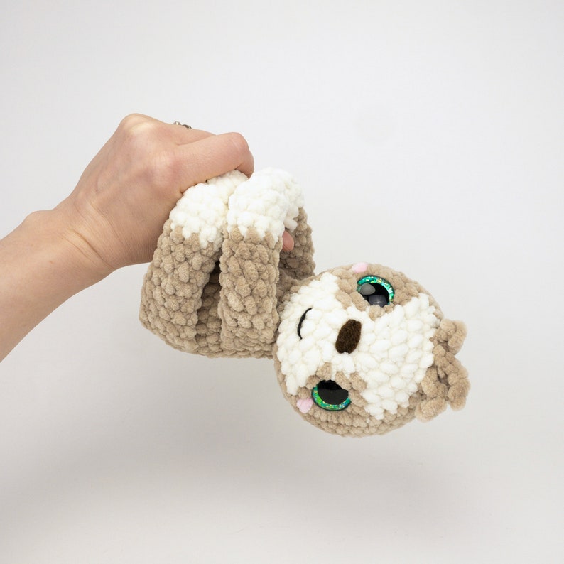 May include: A crocheted sloth plush toy with brown and white fur, green eyes, and a pink nose. The toy is being held by a hand.