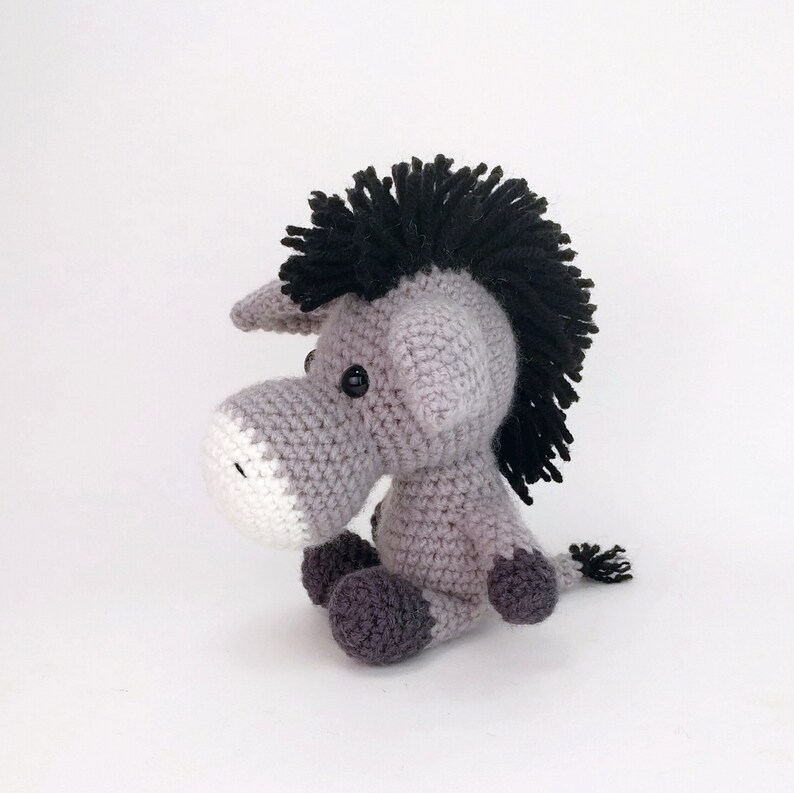 May include: A gray crocheted donkey with black yarn for the mane and tail. The donkey has white accents on its face and black eyes.