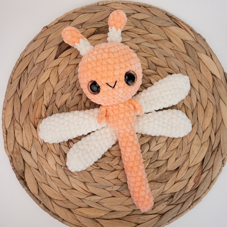May include: A crocheted orange dragonfly toy with white wings. The toy has large black eyes and a smiling mouth.