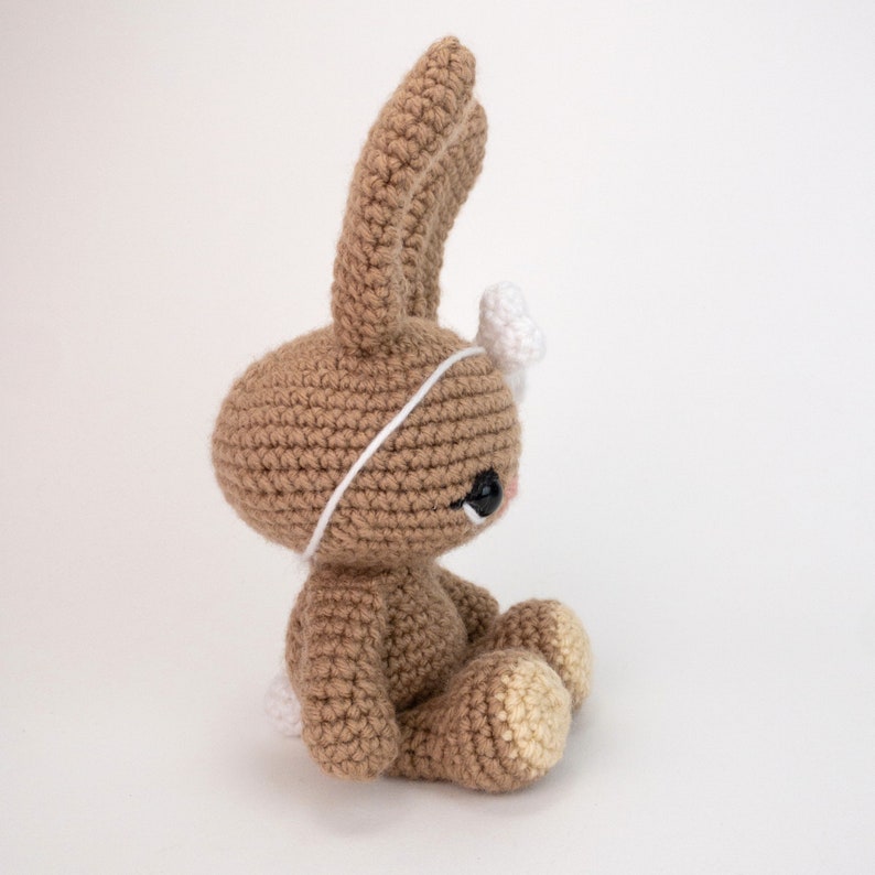 May include: A brown crocheted bunny rabbit with a white crocheted headband and a white bow. The bunny is sitting with its head tilted to the side and its eyes closed.