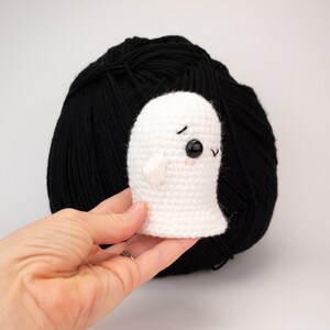 May include: A small, white crochet ghost with black eyes and a pink mouth is held in front of a large ball of black yarn. The ghost has small arms and a friendly expression. The background is plain white.
