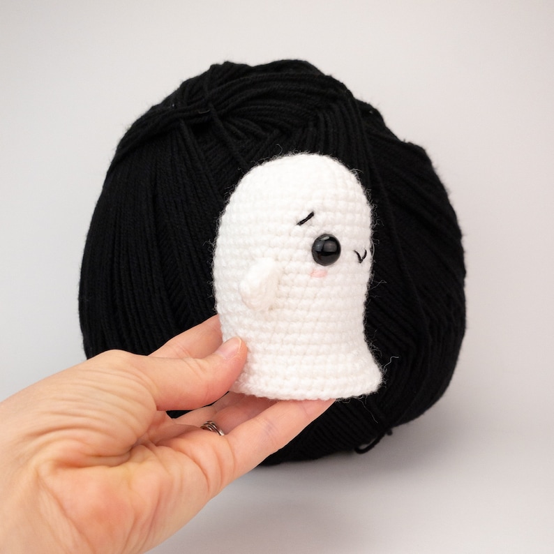 May include: A small, white crochet ghost with black eyes and a pink mouth is held in front of a large ball of black yarn. The ghost has small arms and a friendly expression. The background is plain white.