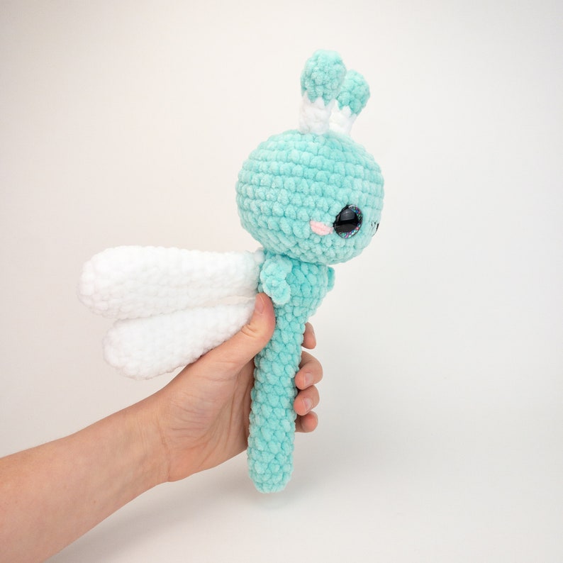 May include: A crocheted dragonfly toy with a light blue body and white wings. The dragonfly has large, black eyes with pink cheeks.