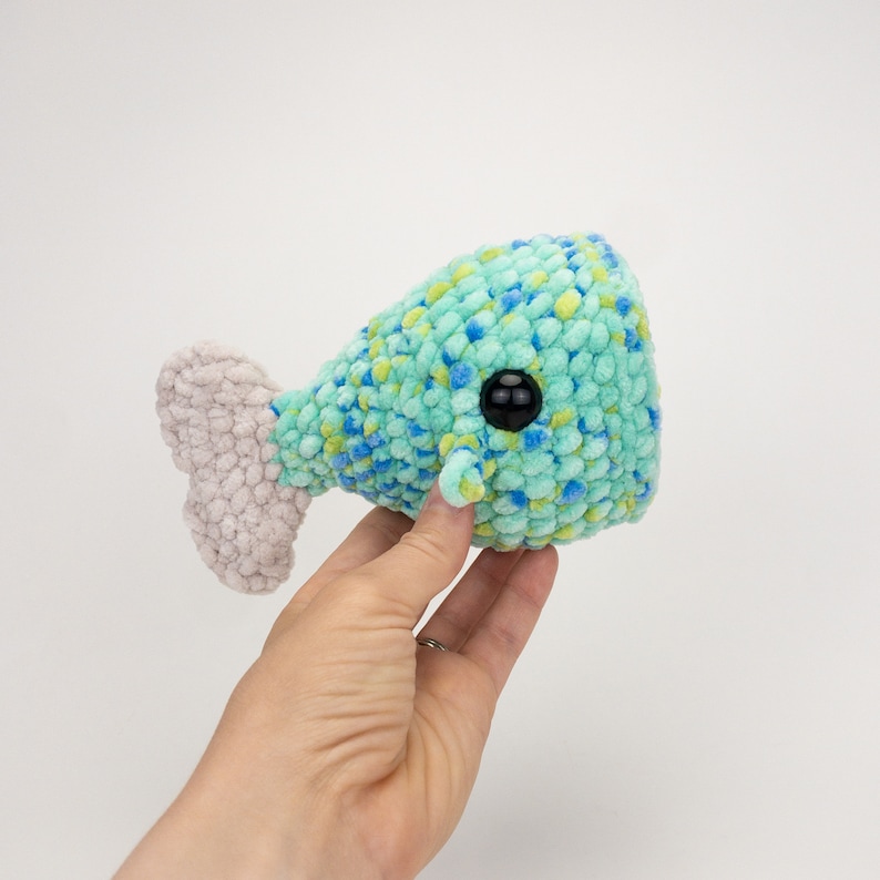 May include: A crocheted whale plush toy with a blue and green speckled body and a white tail fin. The whale has a black button eye.