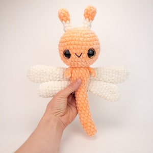 May include: A crocheted orange and white dragonfly plush toy with large black eyes. The toy has a long tail and white wings.