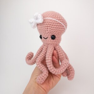 May include: A pink crocheted octopus with a white bow headband. The octopus has eight tentacles and black eyes.
