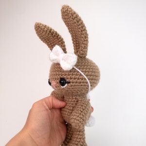 May include: A brown crocheted bunny with a white bow and black eyes. The bunny is standing on a white surface.