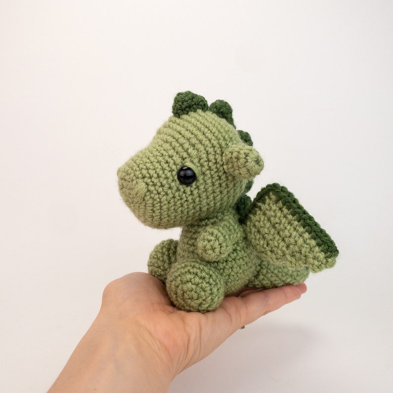 May include: A small green crocheted dragon with black eyes. The dragon is sitting in someone's hand.
