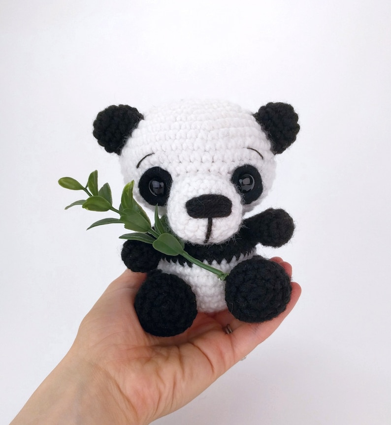 May include: A crocheted panda bear toy holding a green leaf. The panda is white with black ears, eyes, nose, and paws.