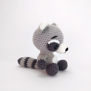 Puede incluir: Un juguete de peluche de mapache tejido a crochet con una cola rayada gris y blanca. El juguete tiene ojos negros y una nariz negra.