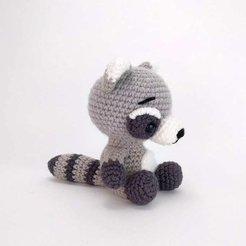 Puede incluir: Un juguete de peluche de mapache tejido a crochet con una cola rayada gris y blanca. El juguete tiene ojos negros y una nariz negra.