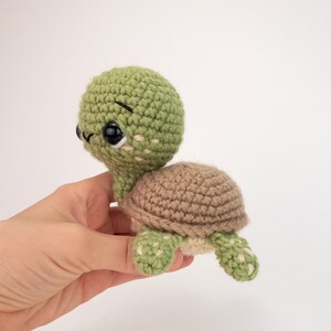 May include: A small crocheted turtle toy with a green shell and brown legs. The turtle has big black eyes and a white face.
