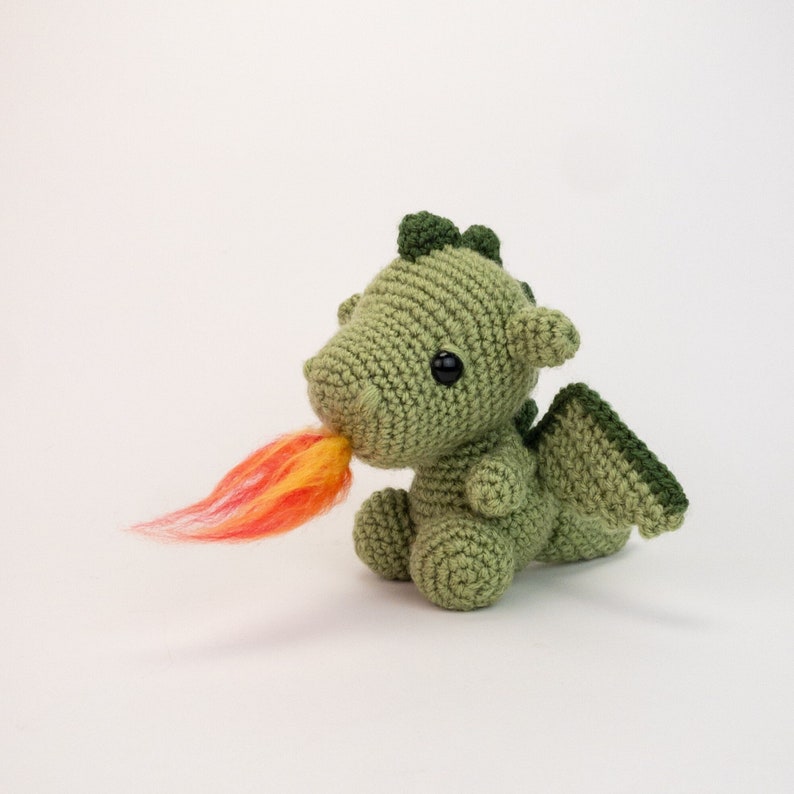 May include: A green crocheted dragon with a red and orange yarn flame coming out of its mouth. The dragon has black eyes and a small, green crocheted wing on its back.