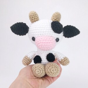 Puede incluir: Un juguete de peluche de vaca tejido a crochet con manchas blancas y negras, nariz rosa y pezu&ntilde;as marrones. La vaca est&aacute; sentada en una mano.