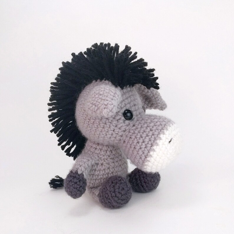 May include: A gray crocheted stuffed animal with a black mohawk, black eyes, and a white snout. The animal is sitting on a white surface.
