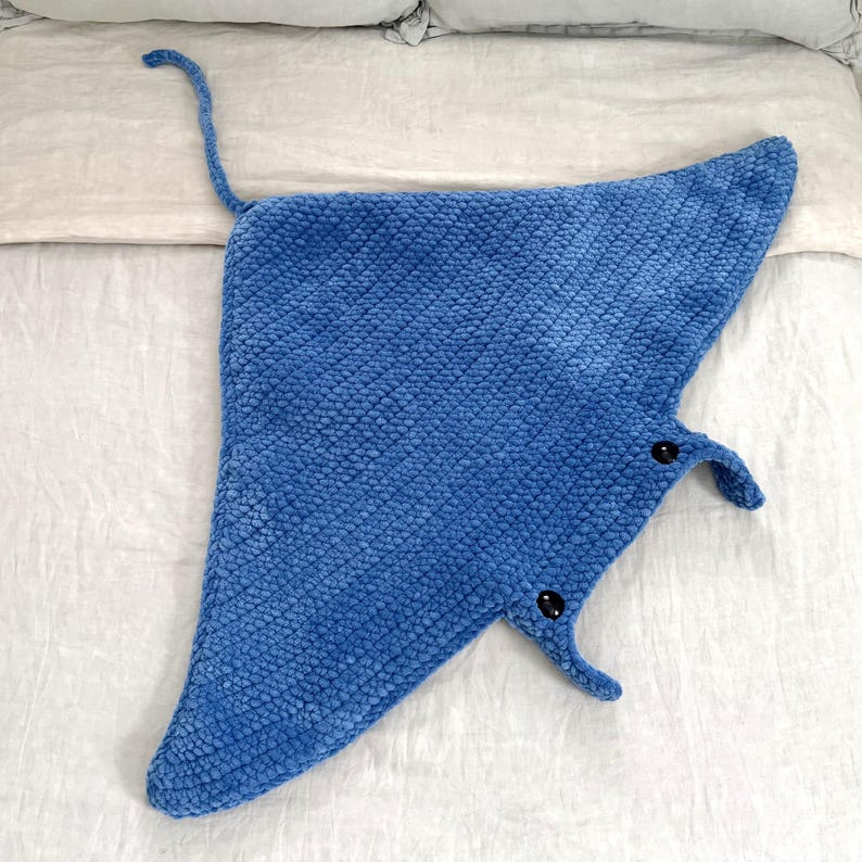 May include: A blue plush manta ray blanket with a tail that can be tied. The blanket has a textured surface and two black buttons for eyes.