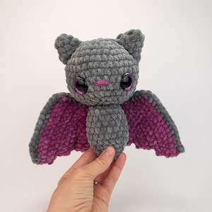 K&ouml;nnte beinhalten: Eine graue geh&auml;kelte Fledermaus mit rosa Fl&uuml;geln. Die Fledermaus hat gro&szlig;e schwarze Augen mit rosa Glitzerakzenten und eine rosa Nase.