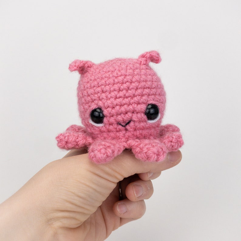 Puede incluir: Un pulpo de crochet rosa con ojos negros y una peque&ntilde;a sonrisa. El pulpo est&aacute; siendo sostenido por una mano.