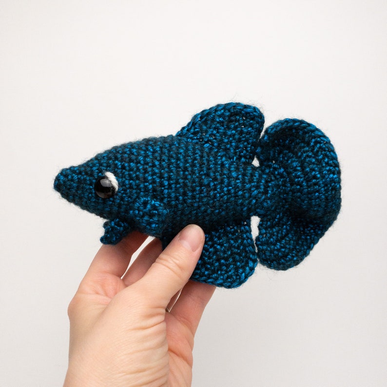 May include: A crocheted blue betta fish toy with a white eye. The fish is being held in a hand.