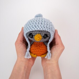 May include: A handmade crochet penguin doll with a light blue hat and ear flaps. The penguin has a gray body, orange belly, and a yellow beak. The doll is held in two hands.