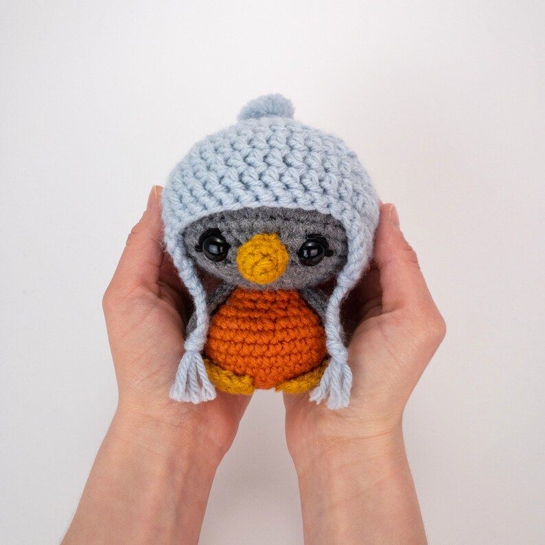 May include: A handmade crochet penguin doll with a light blue hat and ear flaps. The penguin has a gray body, orange belly, and a yellow beak. The doll is held in two hands.