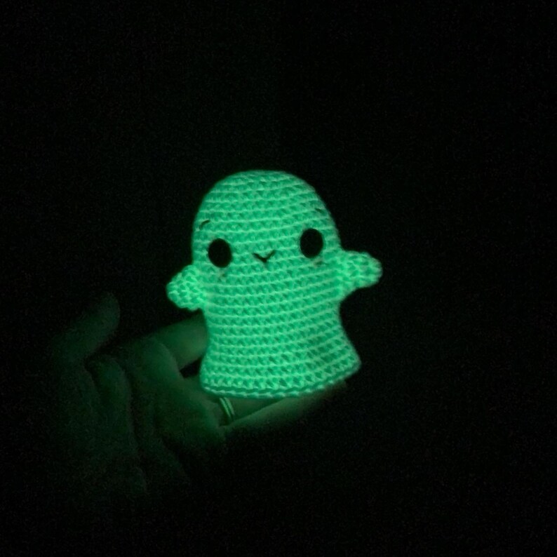 May include: A glowing green crocheted ghost with black eyes and a small smile. The handmade ghost has a rounded body and small arms. The image is set against a dark background, highlighting the bright, luminous quality of the ghost.