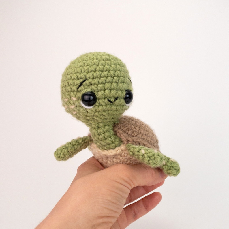 May include: A small crocheted turtle toy with a green shell and brown legs. The turtle has big black eyes and a smiling mouth.