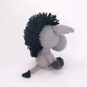 May include: A gray crocheted donkey with a black mane and tail. The donkey is sitting on a white surface.