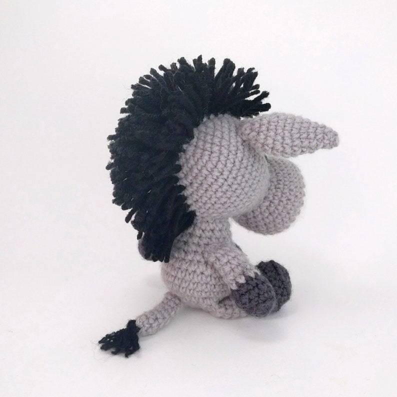 May include: A gray crocheted donkey with a black mane and tail. The donkey is sitting on a white surface.