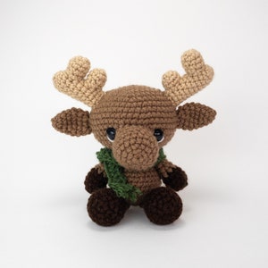 May include: A brown crocheted moose with a green scarf. The moose has large, brown eyes and a small, brown nose. The moose is sitting on a white background.