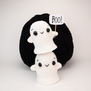 May include: Two white crocheted ghost figures stacked, with black button eyes and rosy cheeks. The top ghost holds a sign saying "BOO!" against a backdrop of black yarn. Halloween-themed decoration.