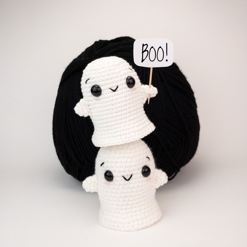May include: Two white crocheted ghost figures stacked, with black button eyes and rosy cheeks. The top ghost holds a sign saying "BOO!" against a backdrop of black yarn. Halloween-themed decoration.