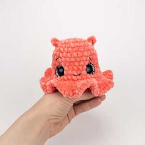 Puede incluir: Un peluche de pulpo de ganchillo de color coral, con ojos grandes y brillantes y una cara sonriente. El peluche tiene un aspecto texturizado y difuso y pequeños tentáculos redondeados. El juguete se sostiene en una mano sobre un fondo blanco.