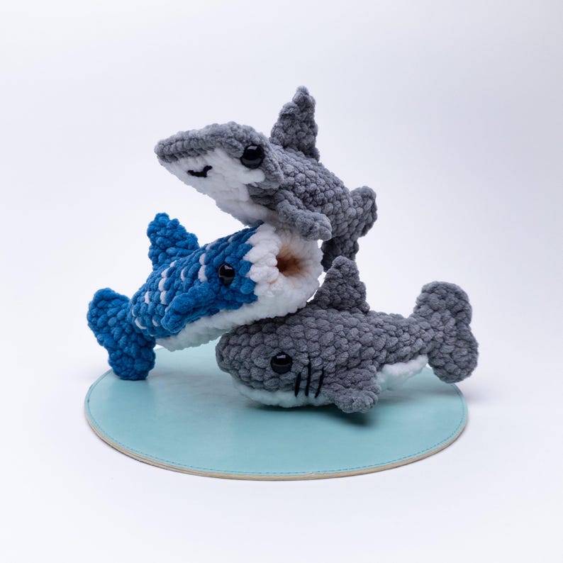 May include: Three crocheted shark plushies stacked on top of each other. The top shark is gray, the middle shark is blue and white, and the bottom shark is gray. The sharks are all facing forward and have black eyes.