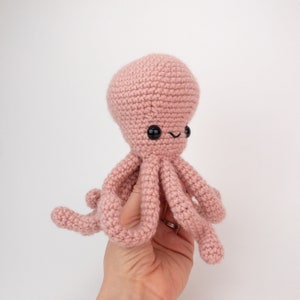 May include: A pink crocheted octopus with black eyes and a smiling mouth. The octopus is being held by a hand.
