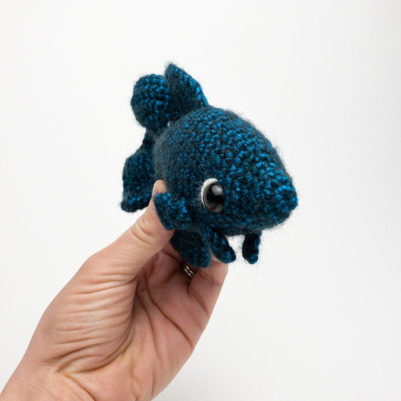 May include: A crocheted blue fish toy with large black eyes. The fish has a fin on its back and two fins on the bottom.