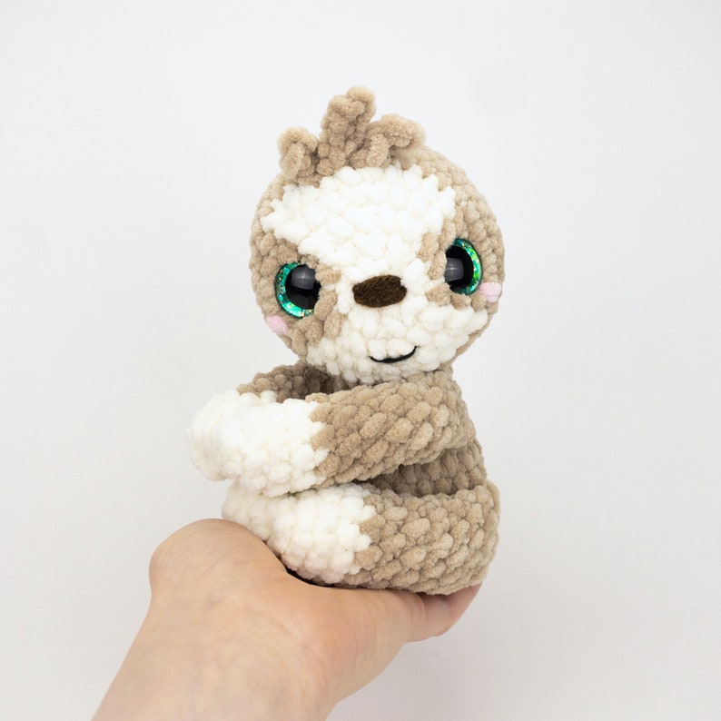 May include: A crocheted sloth plush toy with brown and white fur, green eyes, and a pink nose. The sloth is sitting with its arms crossed.