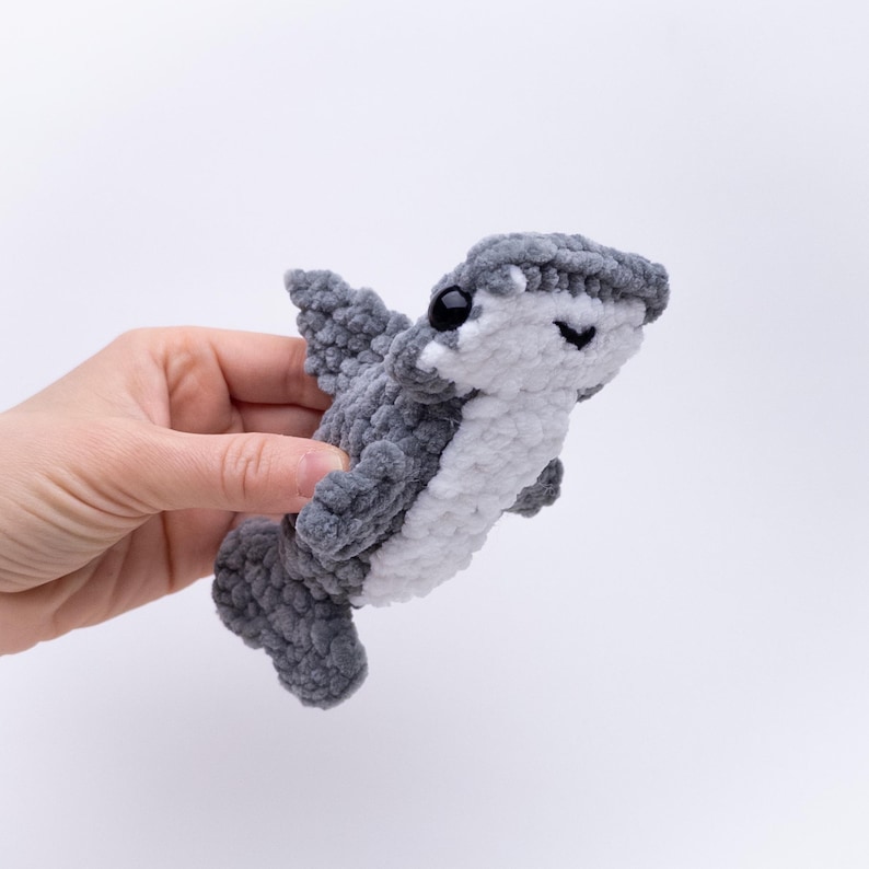 May include: A gray and white crocheted shark plush toy with black eyes. The shark is being held in a hand.