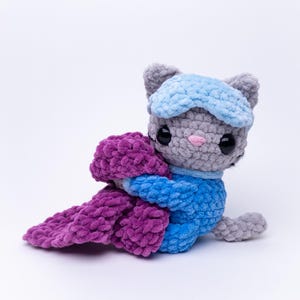 May include: A gray crocheted cat plush toy wearing a light blue sleep mask and a blue outfit. The cat has large black eyes, a small pink nose, and is holding a textured purple crocheted blanket in its arms. The plush is made of soft yarn.