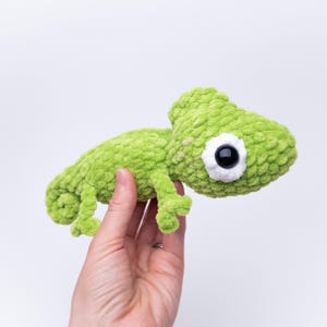 Puede incluir: Un juguete de camaleón verde de peluche hecho a mano, con un ojo grande con borde blanco y una pupila negra. El cuerpo suave y texturizado es de color verde vibrante. El juguete está diseñado para parecerse a un camaleón.