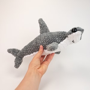 May include: A gray and white crocheted hammerhead shark plush toy with black eyes. The plush toy is being held by a hand.